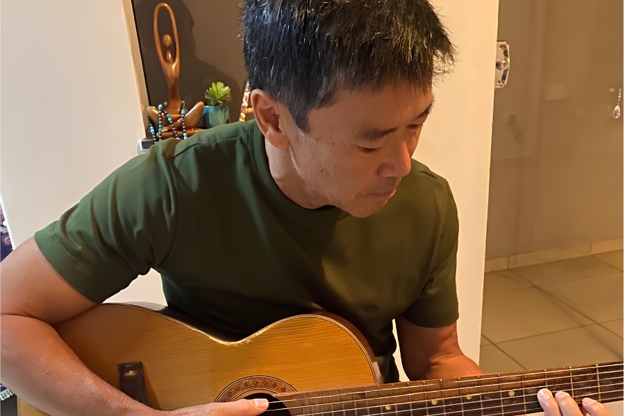Flávio, sentado, tocando violão em casa